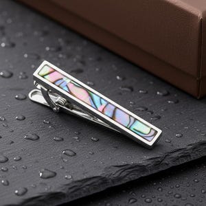 Puede incluir: Un pasador de corbata plateado con una incrustación iridiscente de concha de abulón. El clip rectangular se encuentra sobre una superficie oscura y texturizada con gotas de agua, y una caja marrón en el fondo. Un accesorio elegante.