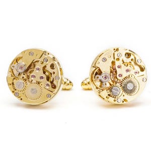 May include: A pair of gold cufflinks with a detailed, intricate design of a watch mechanism.