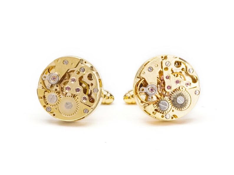May include: A pair of gold cufflinks with a detailed, intricate design of a watch mechanism.
