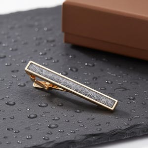 May include: A gold-coloured tie bar featuring a rectangular insert with a grey and white pattern. The tie clip is set against a dark surface with water droplets, with a brown box in the background. Suitable for formal attire.