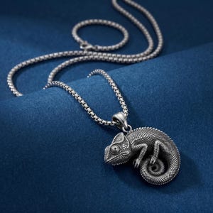 May include: A silver-toned metal pendant shaped like a detailed chameleon with a textured, scaled surface. The pendant is attached to a matching silver-toned box chain necklace, featuring a repeating square link pattern throughout the entire length.