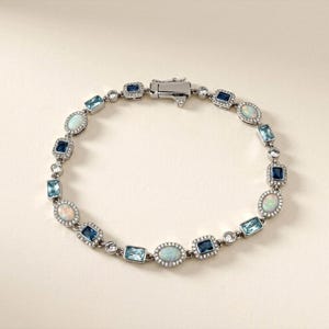 May include: A silver-toned bracelet featuring alternating oval-cut opals and rectangular-cut blue gemstones, including light blue and dark blue varieties. Each stone is surrounded by a halo of small clear crystals, connected by silver links.