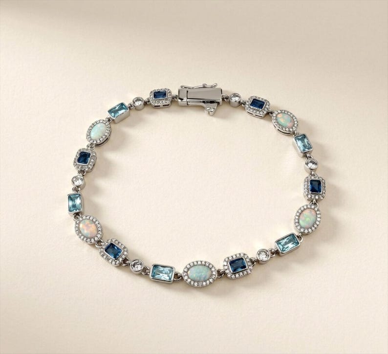 May include: A silver-toned bracelet featuring alternating oval-cut opals and rectangular-cut blue gemstones, including light blue and dark blue varieties. Each stone is surrounded by a halo of small clear crystals, connected by silver links.