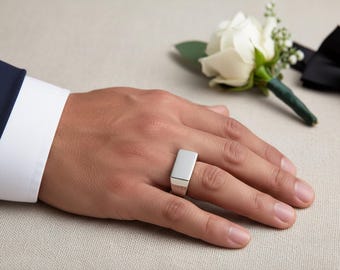 Op de afbeelding: Een zilveren rechthoekige zegelring wordt op een hand gedragen, met een witte rozenknop en een zwarte vlinderdas op de achtergrond. De ring heeft een gepolijste afwerking en een eenvoudig, modern ontwerp.