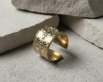 May include: A gold-colored cuff bracelet with an open design. The bracelet features an embossed floral pattern and is adorned with clear, sparkling gemstones. The cuff is set against a neutral, textured background.