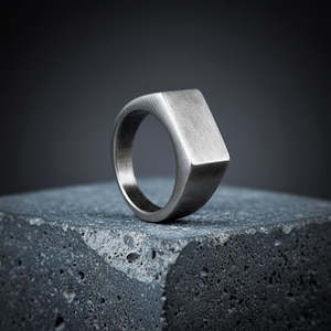 May include: A silver signet ring with a rectangular face. The ring has a brushed metal finish and is displayed on a dark, textured surface. The ring's design is simple and modern. The ring is suitable for everyday wear.