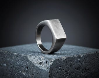 May include: A silver-toned signet ring with a rectangular face. The ring has a brushed metal finish and sits on a dark, textured surface. The ring's design is simple and modern, suitable for everyday wear.