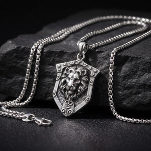 May include: A silver-toned metal pendant necklace featuring a shield-shaped charm with a detailed lion head relief in the center. The pendant is suspended from a silver-toned box chain with a lobster claw clasp, showcasing a polished finish.