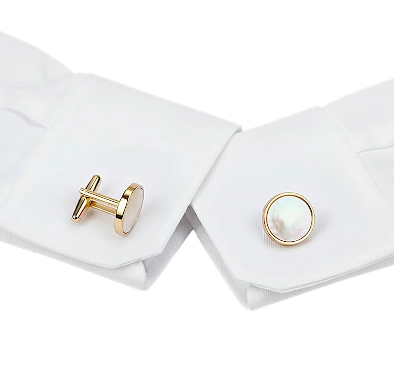 May include: Gold-toned cufflinks with a mother-of-pearl inlay. One cufflink is a bar style, and the other is a round, button style. The cufflinks are displayed on a white dress shirt cuff.