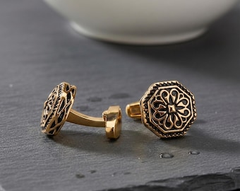 May include: A pair of gold-toned metal cufflinks featuring an octagonal shape with an intricate, openwork floral pattern set against a dark, recessed background. The design includes a textured border and a polished gold finish on the metal posts.