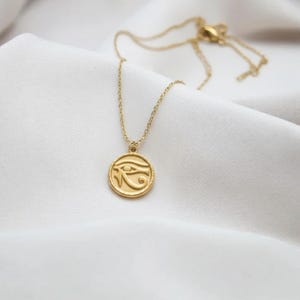 Puede incluir: Un collar dorado con un colgante circular con un diseño de ojo. El collar se muestra sobre una tela blanca texturizada. La cadena y el colgante tienen un acabado brillante, reflejando la luz.