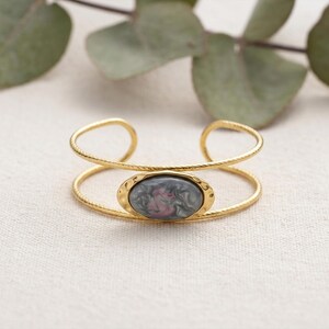 May include: A gold-toned cuff bracelet featuring a double-band design with a twisted texture. The centre of the bracelet is adorned with an oval-shaped cabochon stone displaying a marbled pattern of grey, black, and soft pink swirls.