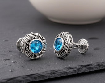 May include: A pair of silver-toned cufflinks featuring an octagonal frame with intricate beaded and rope-like detailing surrounding a central, round, faceted bright blue gemstone. The metal has an oxidized finish to highlight the textured patterns.