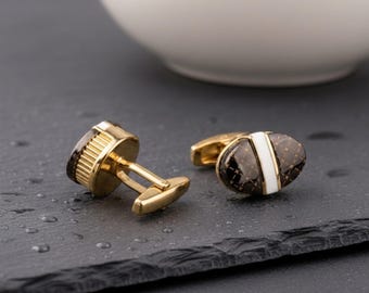 May include: A pair of gold-toned cufflinks featuring an oval-shaped face with a dark brown, grid-patterned inlay and a vertical white stripe down the center. The cufflinks have a polished gold finish and a hinged toggle mechanism on the back.