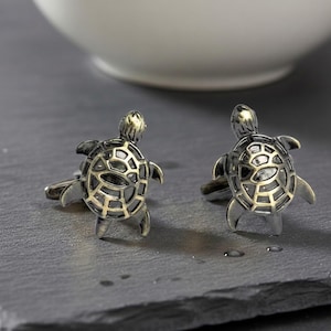 May include: A pair of silver-toned metal cufflinks shaped like sea turtles. Each turtle features a detailed shell with a dark, oxidised finish in the recessed areas and a polished, lighter metallic finish on the raised edges of the shell pattern.
