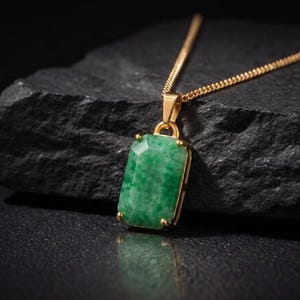 May include: A gold chain necklace featuring a rectangular, faceted green gemstone pendant. The pendant is held by four gold prongs and displays a mottled, semi-translucent texture. The chain is a thin, linked gold style that connects to the top.