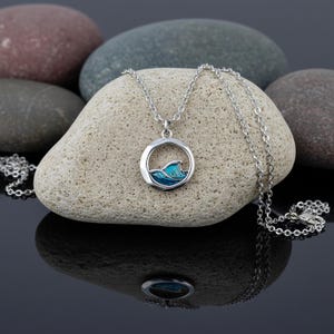Pode incluir: Um colar de prata com um pingente com um desenho de onda azul e turquesa dentro de uma moldura circular. A corrente é delicada e o pingente está sobre uma pedra clara. O colar é uma joia.