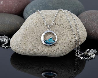May include: A silver necklace with a pendant featuring a blue and turquoise wave design within a circular frame. The chain is delicate, and the pendant is set against a light-colored stone. The necklace is a piece of jewelry.