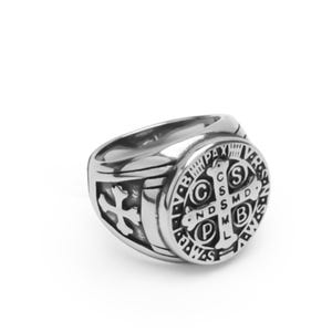 May include: A silver ring with a round, black and silver design featuring a cross and the words "S.C.S.M.D.N.D.S.M.D.B.V.M.L.P.X.V.R.S."