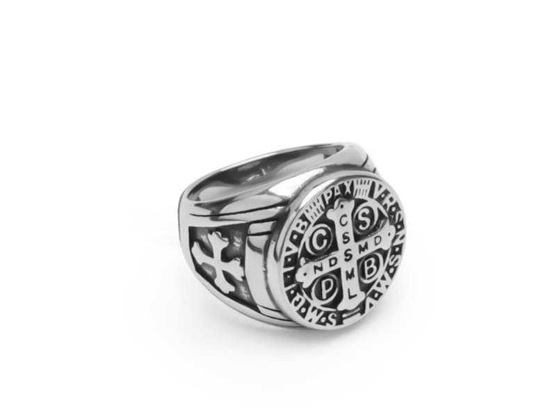 May include: A silver ring with a round, black and silver design featuring a cross and the words "S.C.S.M.D.N.D.S.M.D.B.V.M.L.P.X.V.R.S."