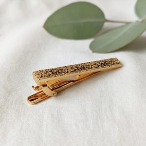 May include: A gold-toned metal tie clip featuring a rectangular front panel with an intricate, dark, engraved scrollwork pattern. The tie clip has a spring-loaded hinge mechanism on the underside, designed to secure a necktie to a dress shirt.