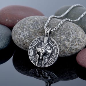 May include: Silver-colored pendant necklace featuring a detailed Spartan helmet design within a circular frame. The pendant hangs from a matching chain, showcasing a classic warrior theme. The necklace is displayed on a reflective surface with gray and brown stones in the background.