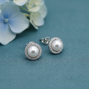 Op de afbeelding: Twee parelstud-oorbellen met een halo van kleine, heldere kristallen. De oorbellen zijn gezet op zilveren stiftjes en worden getoond op een turquoiseblauwe, getextureerde ondergrond, met lichtblauwe en witte bloemen op de achtergrond.