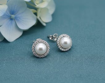May include: Two pearl stud earrings with a halo of small, clear crystals. The earrings are set on a silver post and are displayed on a teal blue textured surface, with light blue and white flowers in the background.