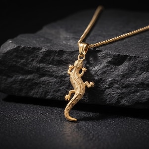 Könnte beinhalten: Eine goldfarbene Metallhalskette mit einem detaillierten Eidechsenanhänger. Der Anhänger ist als kleine Eidechse mit strukturierter Haut und geschwungenem Schwanz geformt und hängt an einer passenden goldfarbenen Kette am Kopf.