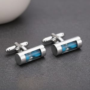 May include: A pair of silver-toned metal cufflinks featuring a cylindrical design with a clear glass window revealing bright blue sand inside. Each cufflink has a standard hinged toggle closure mechanism attached to the back of the cylinder.