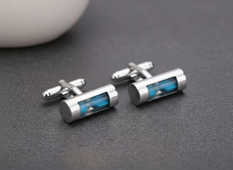 May include: A pair of silver-toned metal cufflinks featuring a cylindrical design with a clear glass window revealing bright blue sand inside. Each cufflink has a standard hinged toggle closure mechanism attached to the back of the cylinder.