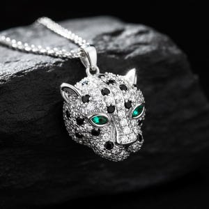 Puede incluir: Un collar con colgante plateado que representa una cabeza de leopardo. El colgante está adornado con piedras preciosas claras y negras, con ojos verde esmeralda. El collar se muestra sobre una superficie oscura y texturizada.