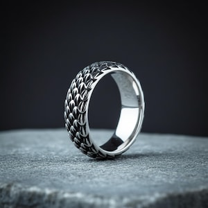 Peut inclure: Une bague argentée avec un motif d'écailles détaillé sur le pourtour. La bague est mise en valeur sur un fond sombre, soulignant son design complexe et son éclat métallique. C'est un bijou.