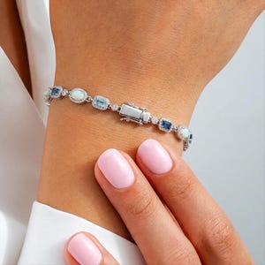 May include: A silver-toned bracelet featuring alternating oval-shaped opals and rectangular-cut blue gemstones, each surrounded by small clear stones. The bracelet includes a rectangular metal clasp and is constructed with a delicate chain link design.