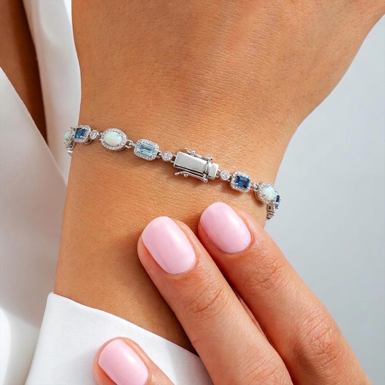 May include: A silver-toned bracelet featuring alternating oval-shaped opals and rectangular-cut blue gemstones, each surrounded by small clear stones. The bracelet includes a rectangular metal clasp and is constructed with a delicate chain link design.