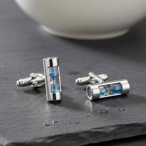 May include: A pair of silver-toned metal cufflinks, each featuring a miniature cylindrical hourglass containing blue sand. The cufflinks have a polished finish and a standard toggle closure mechanism, designed to be worn on shirt cuffs.
