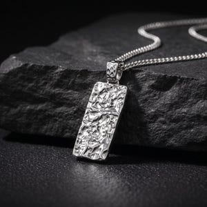May include: A silver-toned rectangular pendant with a highly textured, hammered surface suspended from a matching silver-toned curb chain. The pendant features an irregular, cratered finish that reflects light across its metallic surface.