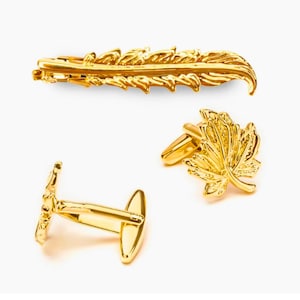 Könnte beinhalten: Ein Set goldfarbener Accessoires, darunter eine Krawattenklammer und zwei Paar Manschettenknöpfe. Die Krawattenklammer hat ein Federdesign, während ein Paar Manschettenknöpfe ein Blattdesign aufweist.