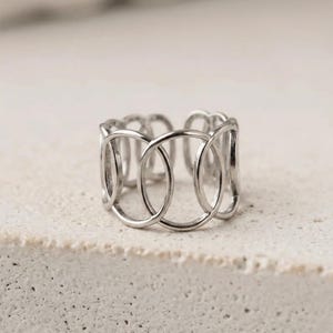 Op de afbeelding: Een zilverkleurige ring met een opengewerkt ontwerp bestaande uit een reeks onderling verbonden ovale schakels. De band heeft een doorlopend, herhalend patroon van gladde, gepolijste metalen lussen die een brede, luchtige structuur vormen.