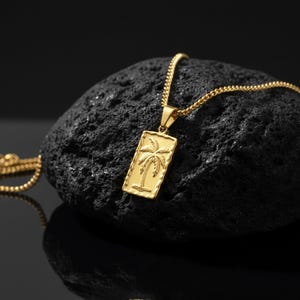 Puede incluir: Un collar dorado con un colgante rectangular con un diseño de palmera. El colgante cuelga de una cadena dorada a juego. El collar se exhibe sobre una superficie oscura y texturizada, creando un contraste con la brillante joyería.
