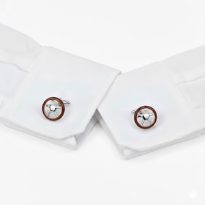 May include: A pair of silver cufflinks with a brown circular border and a steering wheel design. The cufflinks are attached to the white cuffs of a dress shirt, creating a classic and elegant look.