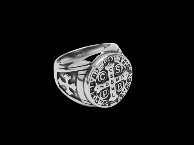 May include: A silver ring with a round, black, detailed design featuring a cross and the words "S.P.D.  ND.S.M.D.  C.S.S.M.D.  P.L.  V.B.  PAX  VBS".