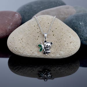 Op de afbeelding: Een zilverkleurige panda beer hanger ketting. De panda houdt een bamboestengel vast met groene edelsteenbladeren. De ketting wordt getoond op een lichtgekleurde steen met een reflecterend oppervlak.