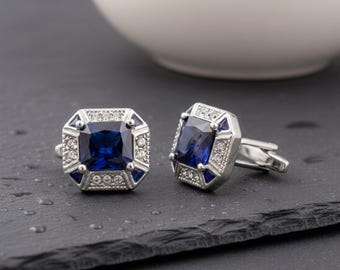 May include: A pair of silver-toned cufflinks featuring a square-cut deep blue center stone. The frame is accented with smaller clear white crystals and triangular blue stones set in a geometric pattern, creating a polished and structured appearance.