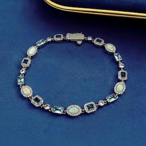 May include: A silver-toned bracelet featuring alternating oval-cut opals and rectangular-cut blue gemstones. Each stone is surrounded by a halo of small, clear pav&eacute;-set crystals. The bracelet includes a secure metal box clasp closure mechanism.