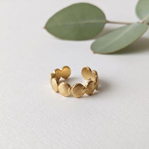 Può includere: Un anello aperto color oro caratterizzato da un motivo ripetitivo di piccole conchiglie testurizzate collegate tra loro in una fascia circolare. Il metallo ha una finitura lucida e il design è composto da diverse forme di conchiglie singole.