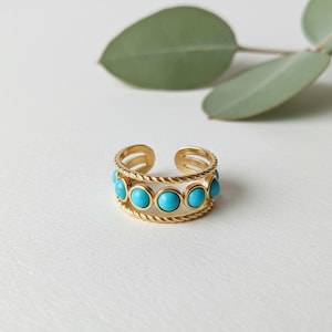 Op de afbeelding: Goudkleurige verstelbare ring met een rij ronde, turquoise gekleurde stenen. De ring heeft een gedraaid touwontwerp rond de stenen en een gespleten schacht. De ring staat tegen een witte achtergrond met een groen blad.
