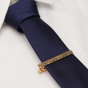 May include: A navy blue tie with a gold tie clip. The tie clip has a textured design.