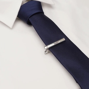 May include: A navy blue tie with a silver tie bar, set against a white dress shirt. The tie bar is rectangular and has a clasp to secure it to the tie. The tie is made of a smooth fabric.