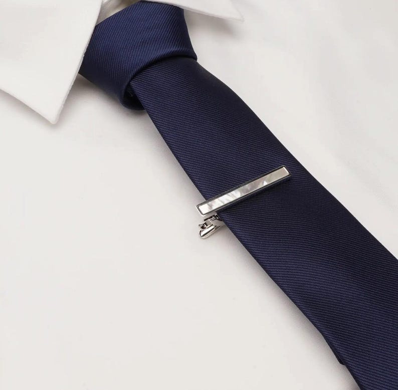 May include: A navy blue tie with a silver tie bar, set against a white dress shirt. The tie bar is rectangular and has a clasp to secure it to the tie. The tie is made of a smooth fabric.